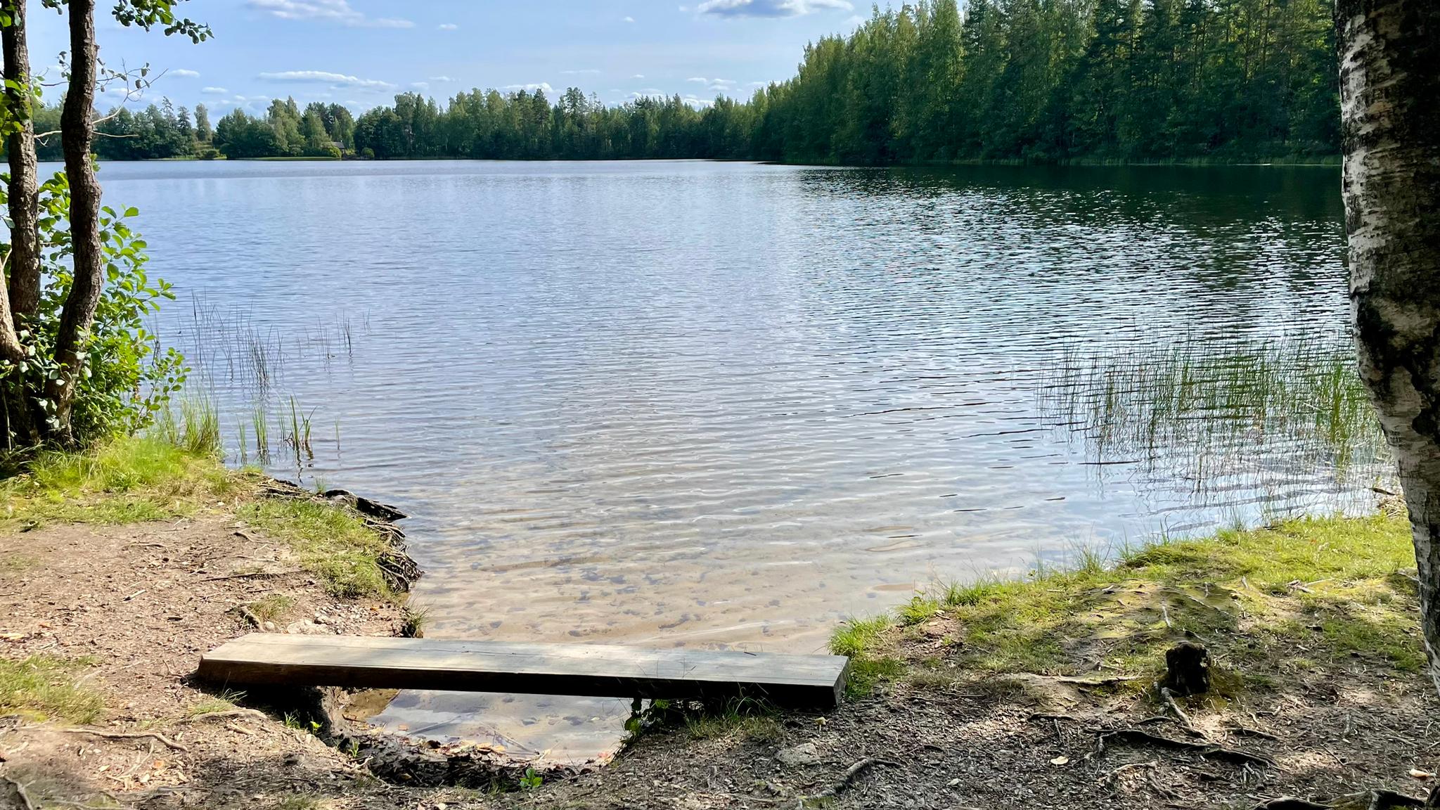 A lake with clear water. The sand at the bottom is clearly visible through the water. There is a small cove with a plank.