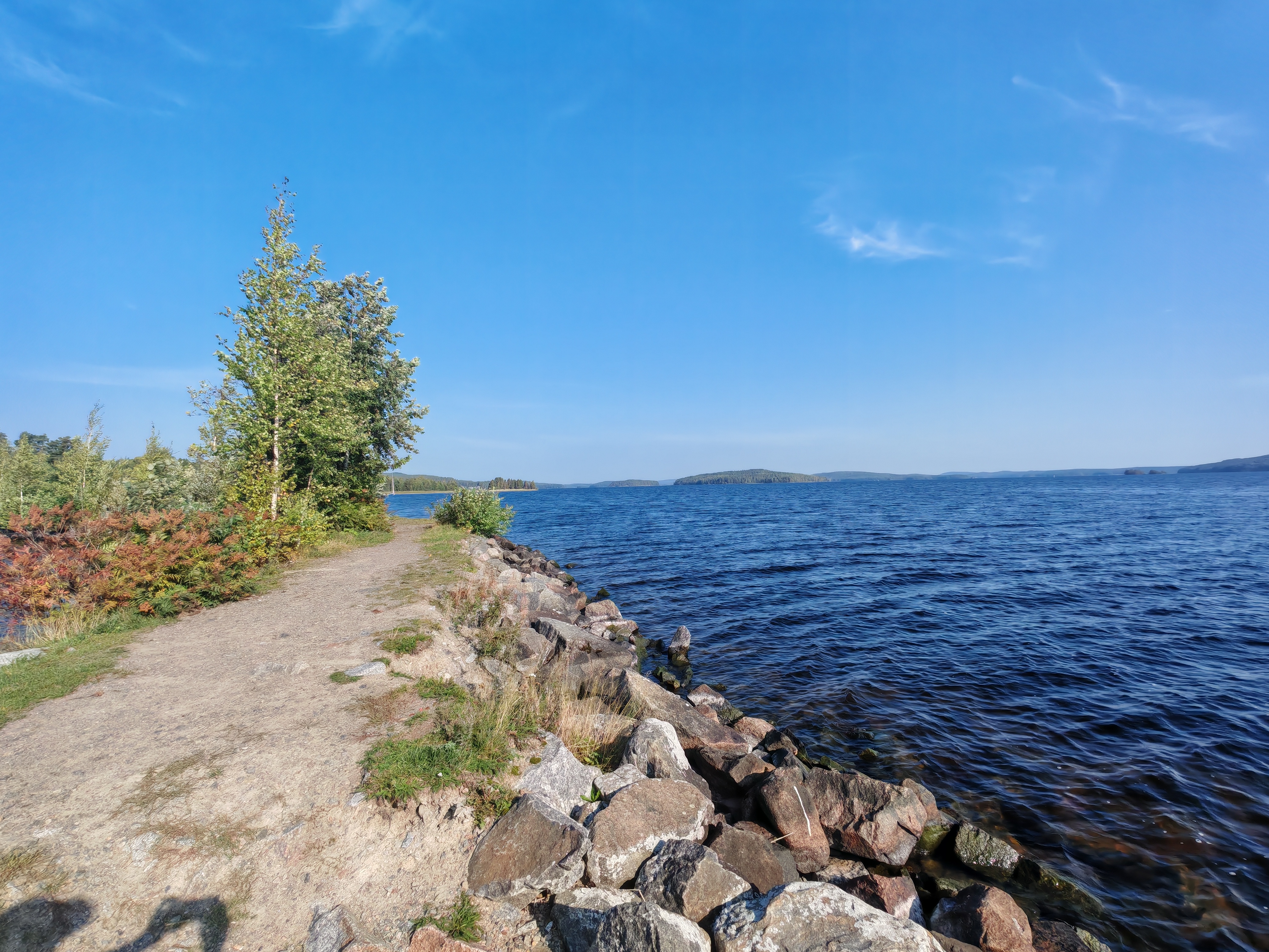 Kuvassa näkyy hiekkapolku. Oikealla puolella polkua on sinisenä kimmeltävä järvi, jonka rannassa on kivistä rakennettu penger.