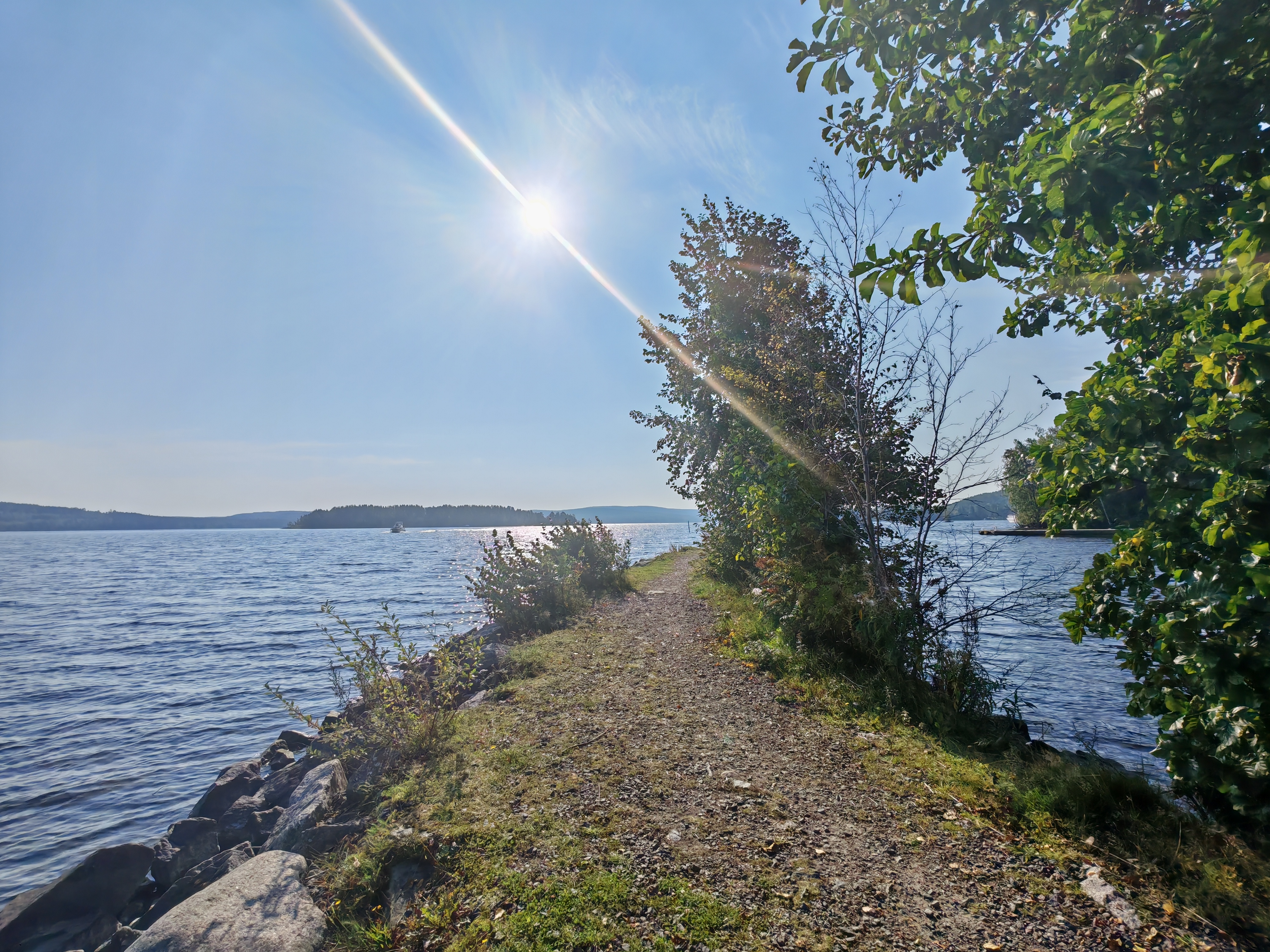 Kuvassa näkyy kapea polku, joka kulkee aivan Päijänteen rannassa. Vasemmalla puolella avautuu kirkasvetinen järvi, jonka pinta kimmeltää auringonvalossa. Oikealla puolella polkua reunustavat vihreät puut ja pensaat, jotka luovat varjoisaa kontrastia avoimelle järvimaisemalle. Taustalla näkyy kauempana järven selällä pieni saari sekä horisonttia kehystävät metsät. Aurinko paistaa korkealla kirkkaalta taivaalta, luoden kuvan tunnelmaan kesäisen lämpimän ja rauhallisen sävyn.