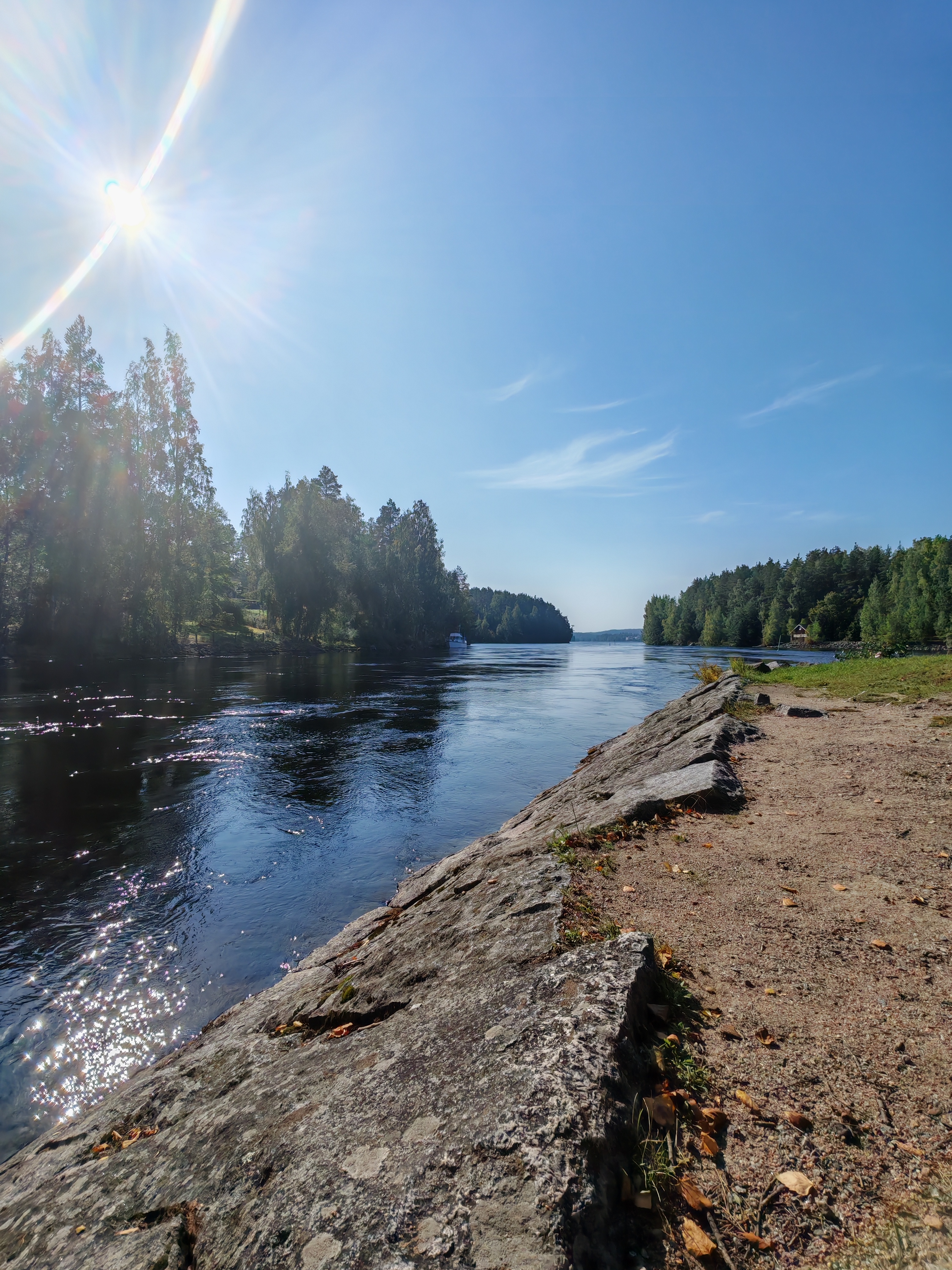 Kuvassa näkyy kirkas ja aurinkoinen jokimaisema, jossa veden pinta kimmeltää auringonvalossa. Joen varrella on kivinen ranta, joka sopii hyvin rauhalliseen kalastukseen tai rentoutumiseen luonnon äärellä.