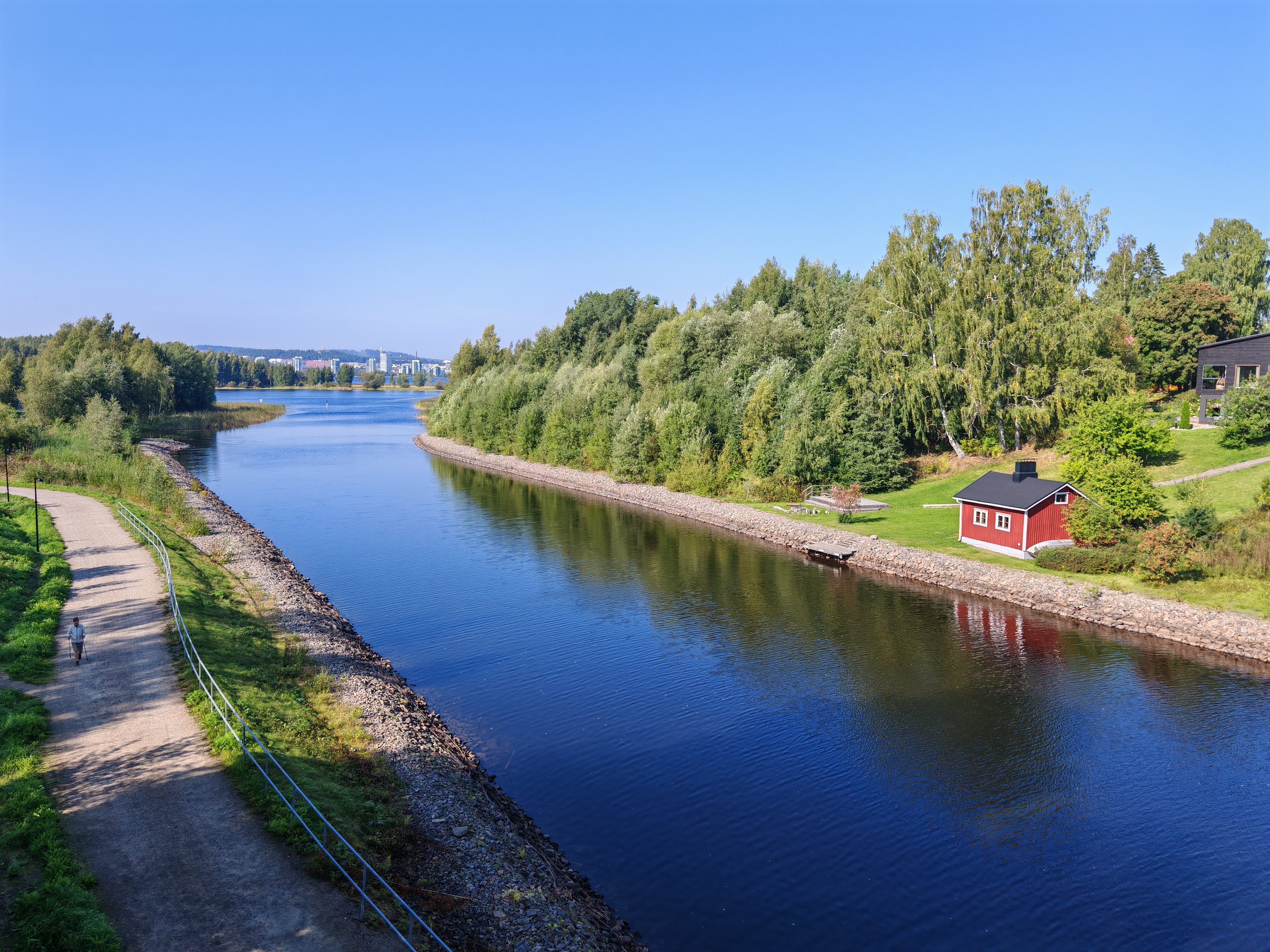 Kuvassa näkyy rauhallinen Äijälänsalmi, jota reunustavat vehreät rannat ja metsikkö. Salmi yhdistää Jyväsjärven Päijänteeseen. Vasemmalla rannalla kulkee hiekkatie, jolla kävelee yksi henkilö, ja oikealla rannalla on punainen pieni mökki. Taustalla avautuu järvimaisema, jonka horisontissa siintävät kaupungin rakennukset ja sininen taivas.