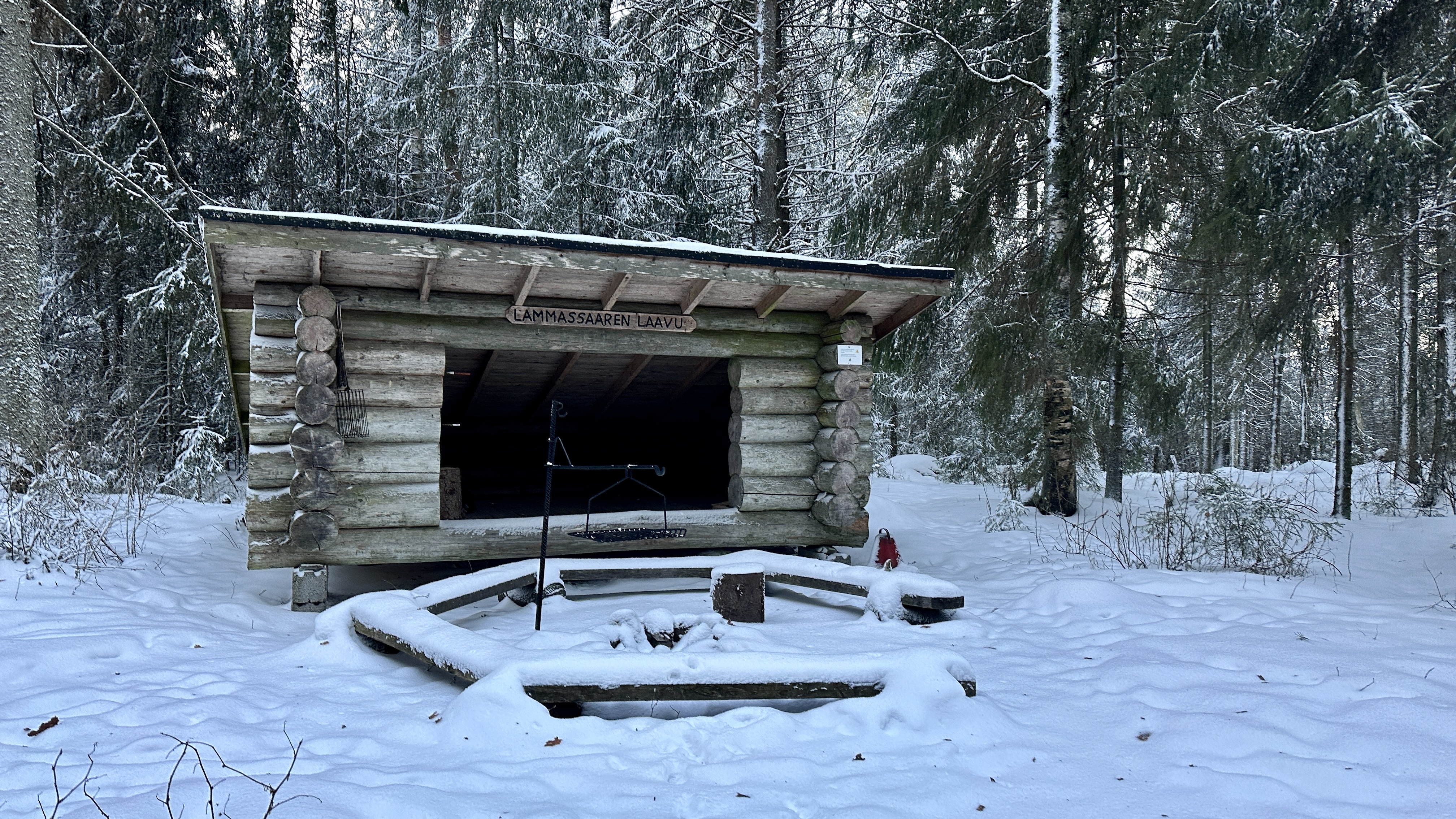 In the winter forest there is a lean-to shelter with a campfire site in front of it, covered in snow.