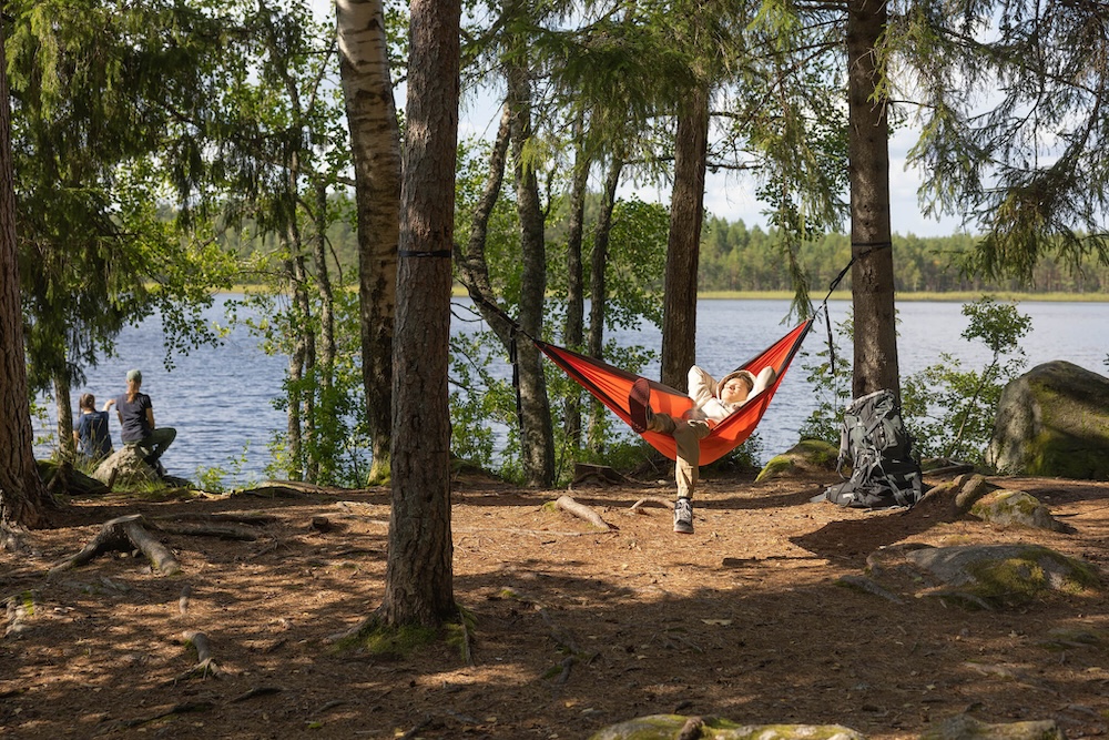 Mies rentoutuu riippumatossa järven rannalla metsässä.