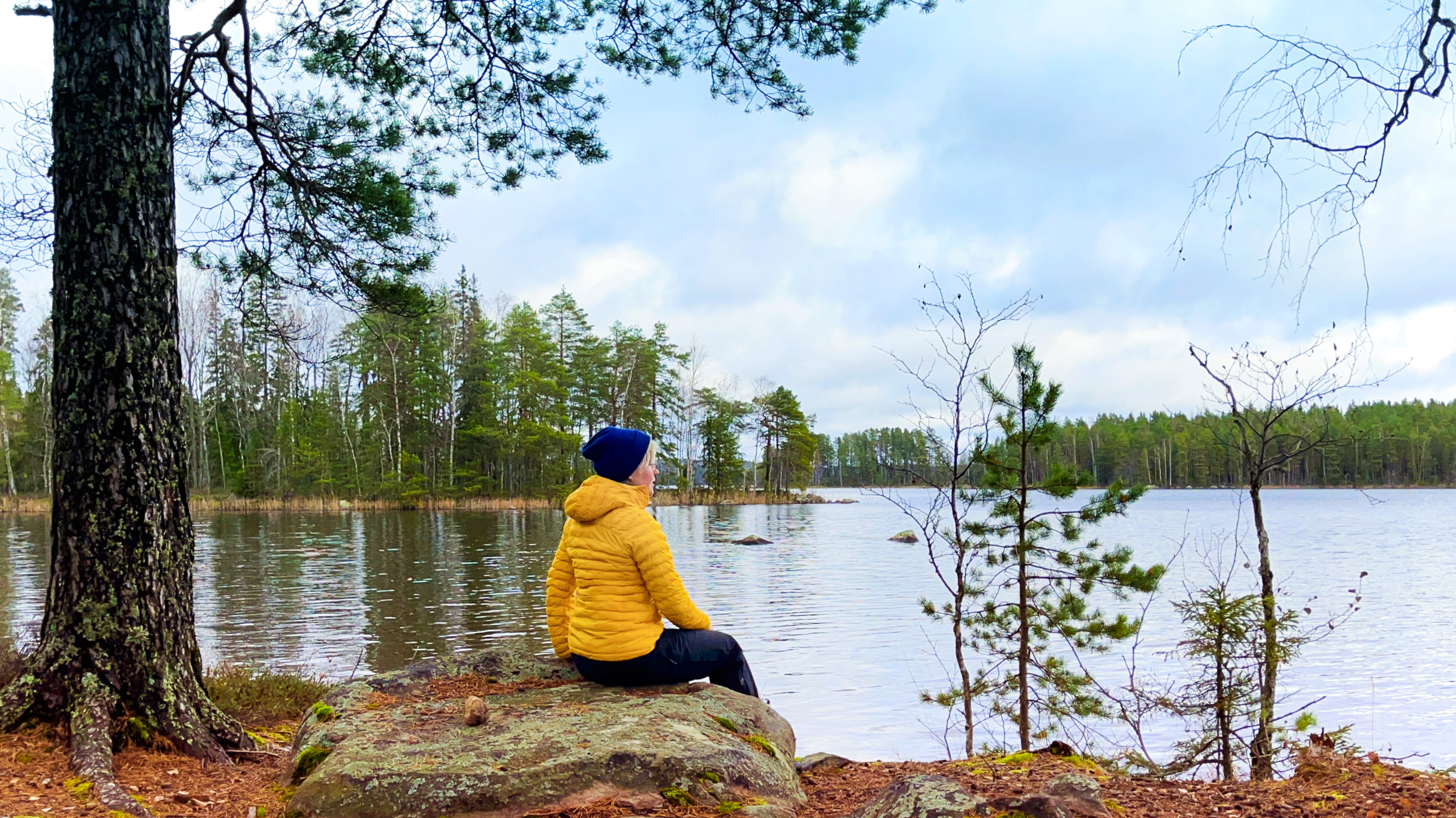 A person wearing a yellow jacket and a blue beanie sits on a rock by the shore of Kivijärvi, with a forested lake view in the background.