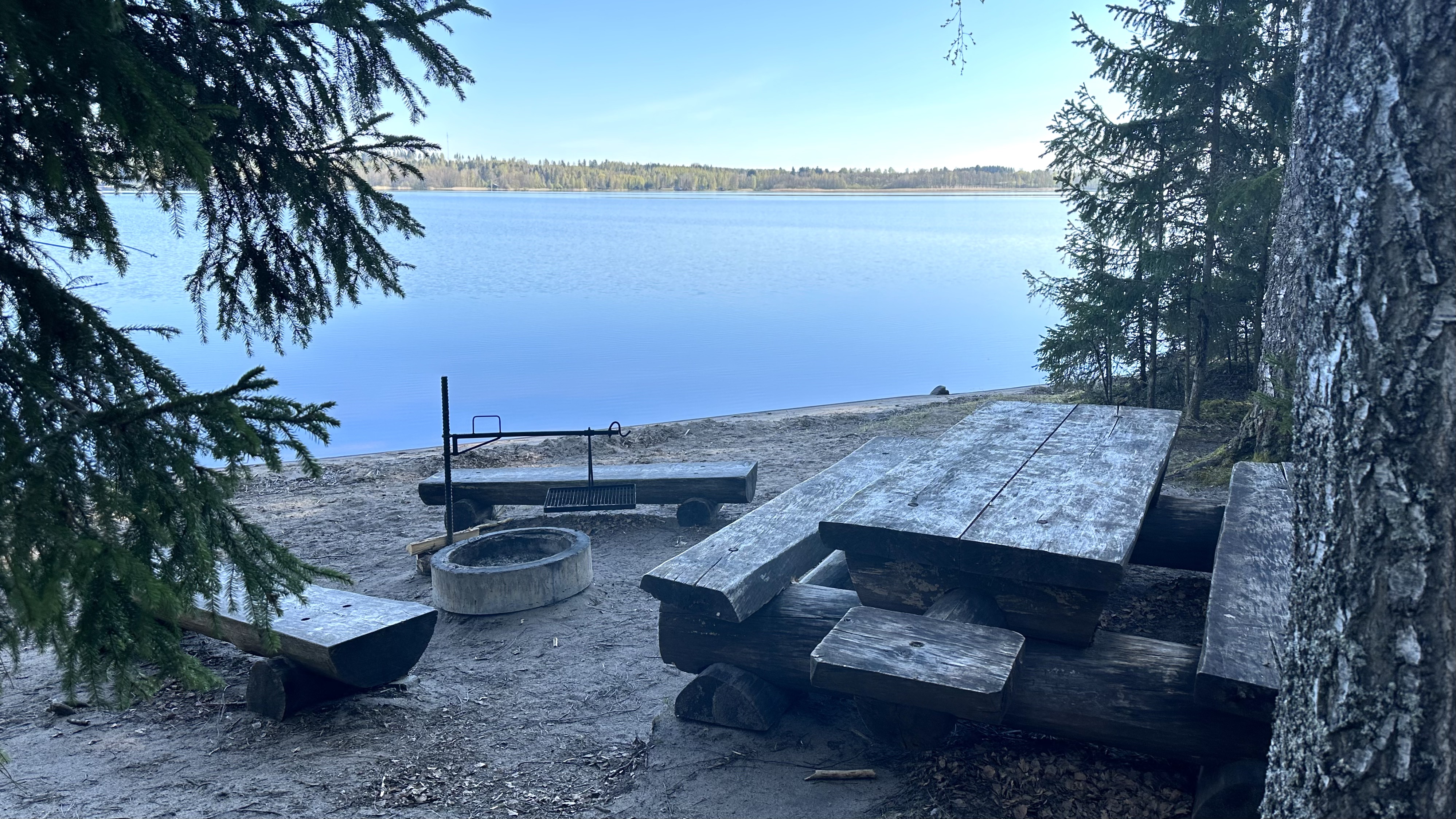 By the lake there is a wooden table with benches, two wooden benches and a campfire site.