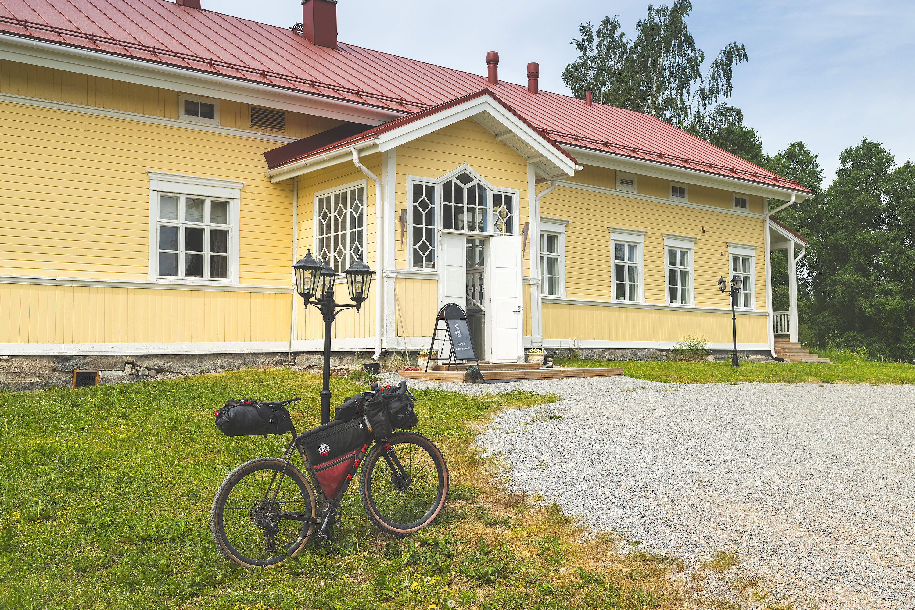 A yellow wooden farmhouse in summer. A gravel bike leaning to a light post infront.