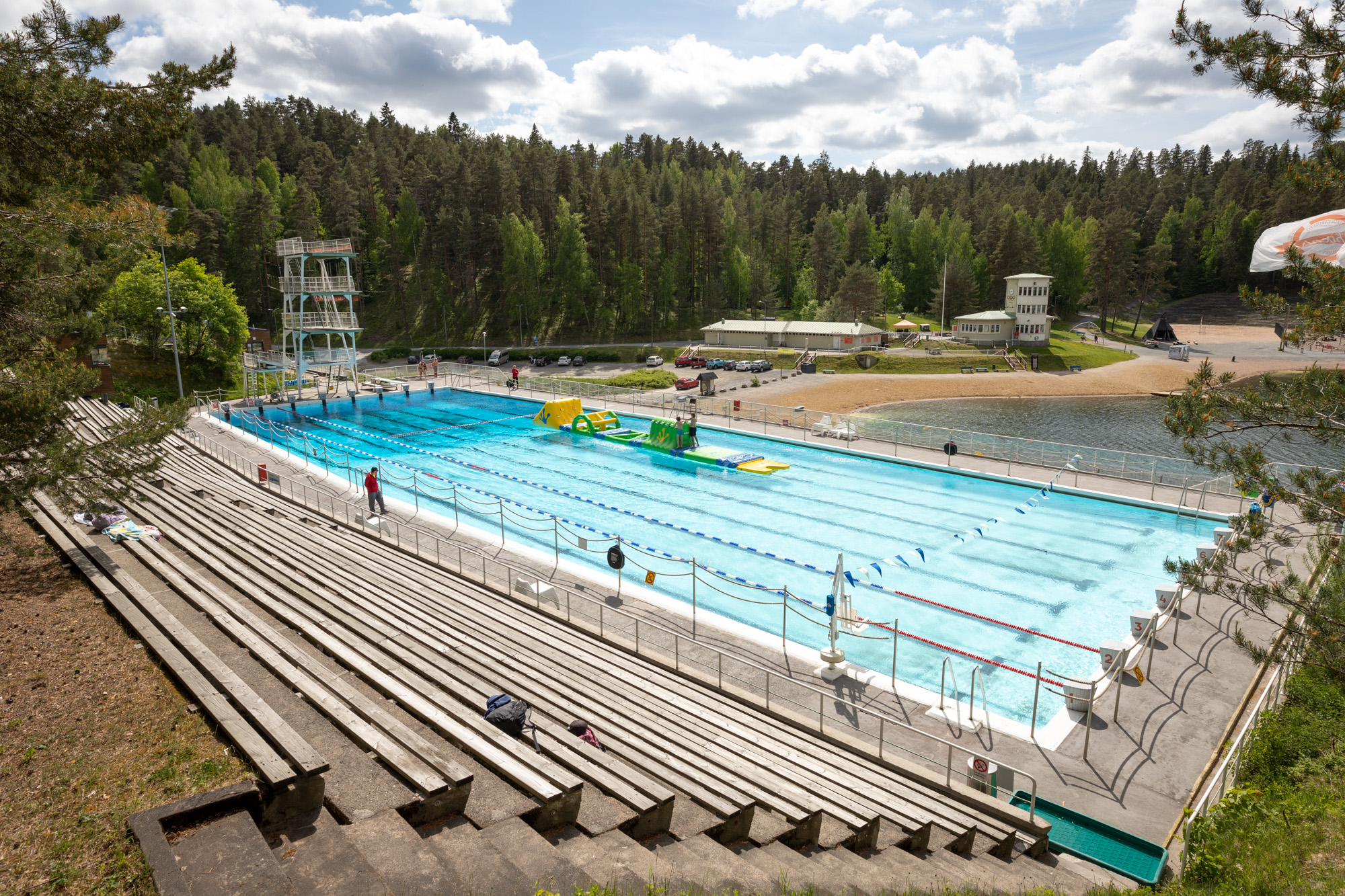 Edessä maauimalan katsomo ja uima-allas. Taustalla hiekkainen uimaranta ja kahvilarakennus