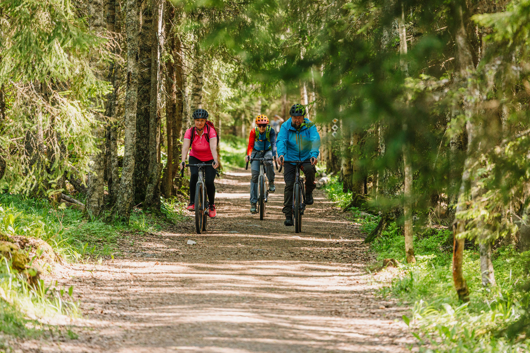 Kolme pyöräilijää pyöräilee metsän reunustamalla hiekkatiellä.