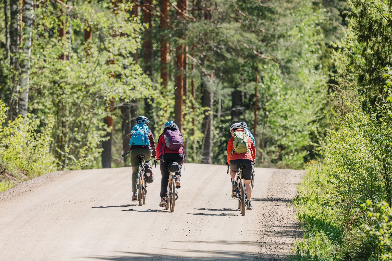 Kolme pyöräilijää kulkevat hiekkatiellä pyöräillen ylämäkeen.