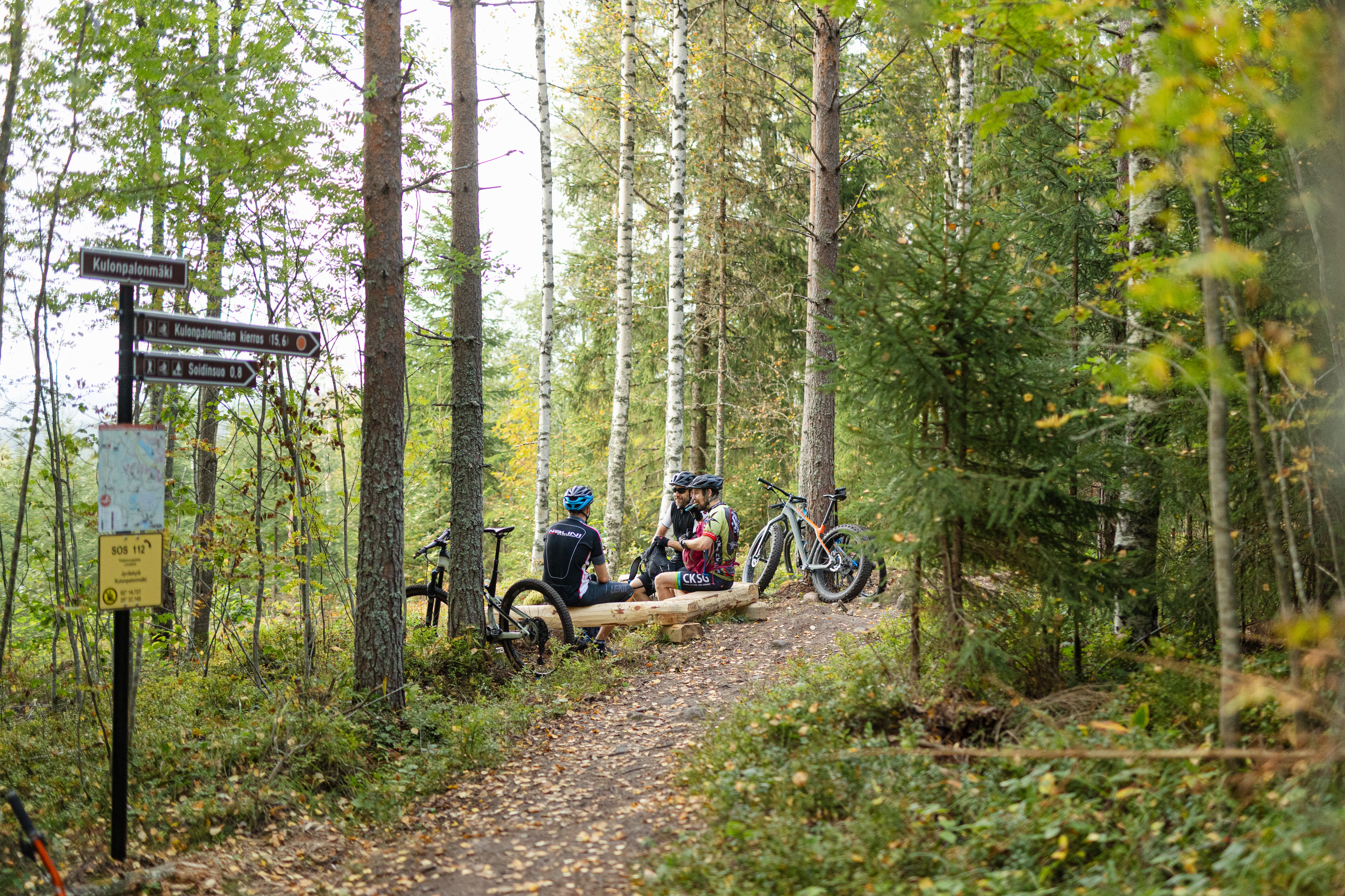 Etualalla opastepylväs. Taaempana 3 henkilöä istumassa polun varren taukopaikalla maastopyörien kanssa.