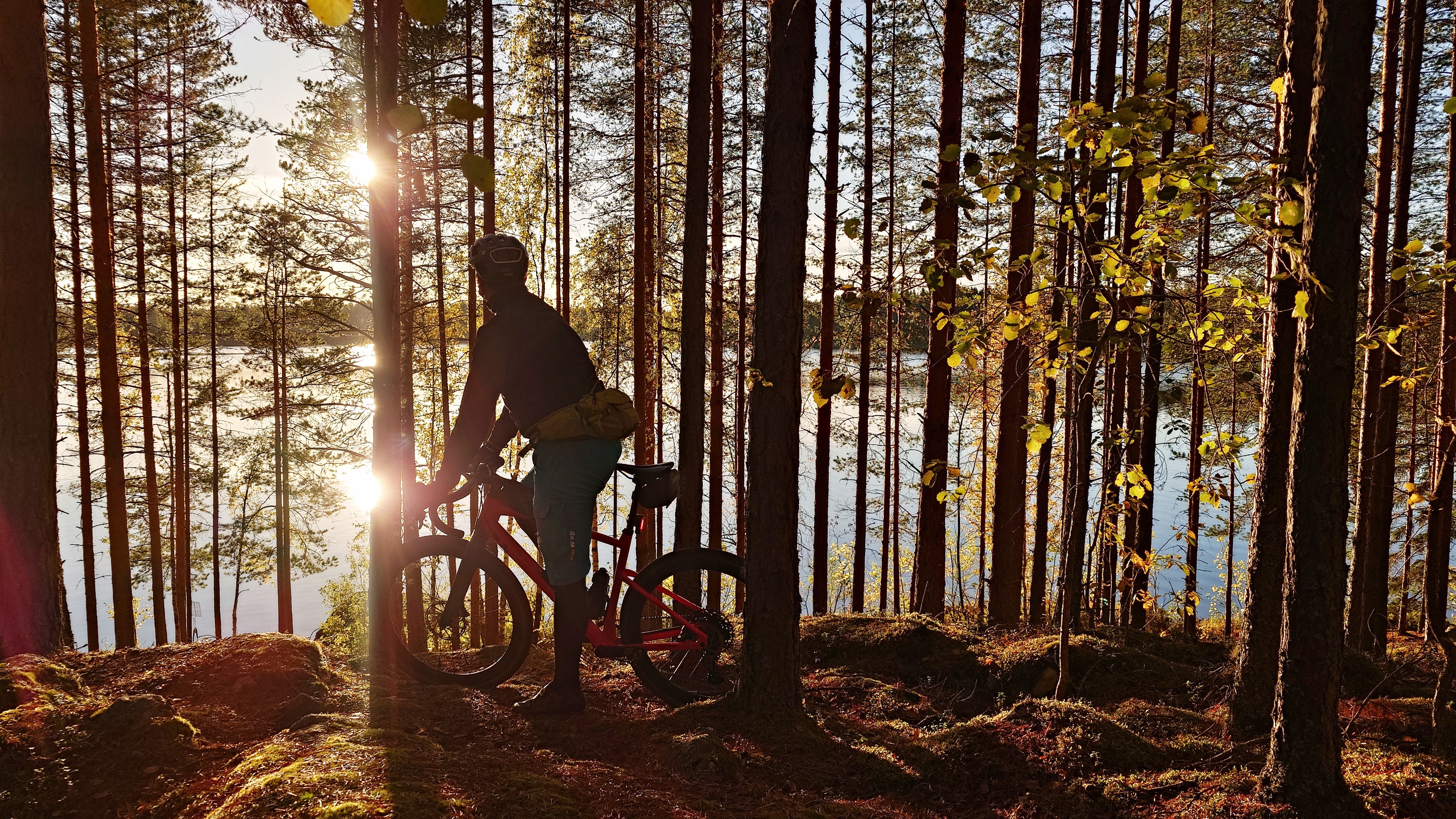 Pyöräilijä metsässä kivikossa, aurinko paistaa ja heijastuu metsän takana olevaan järven pintaan.