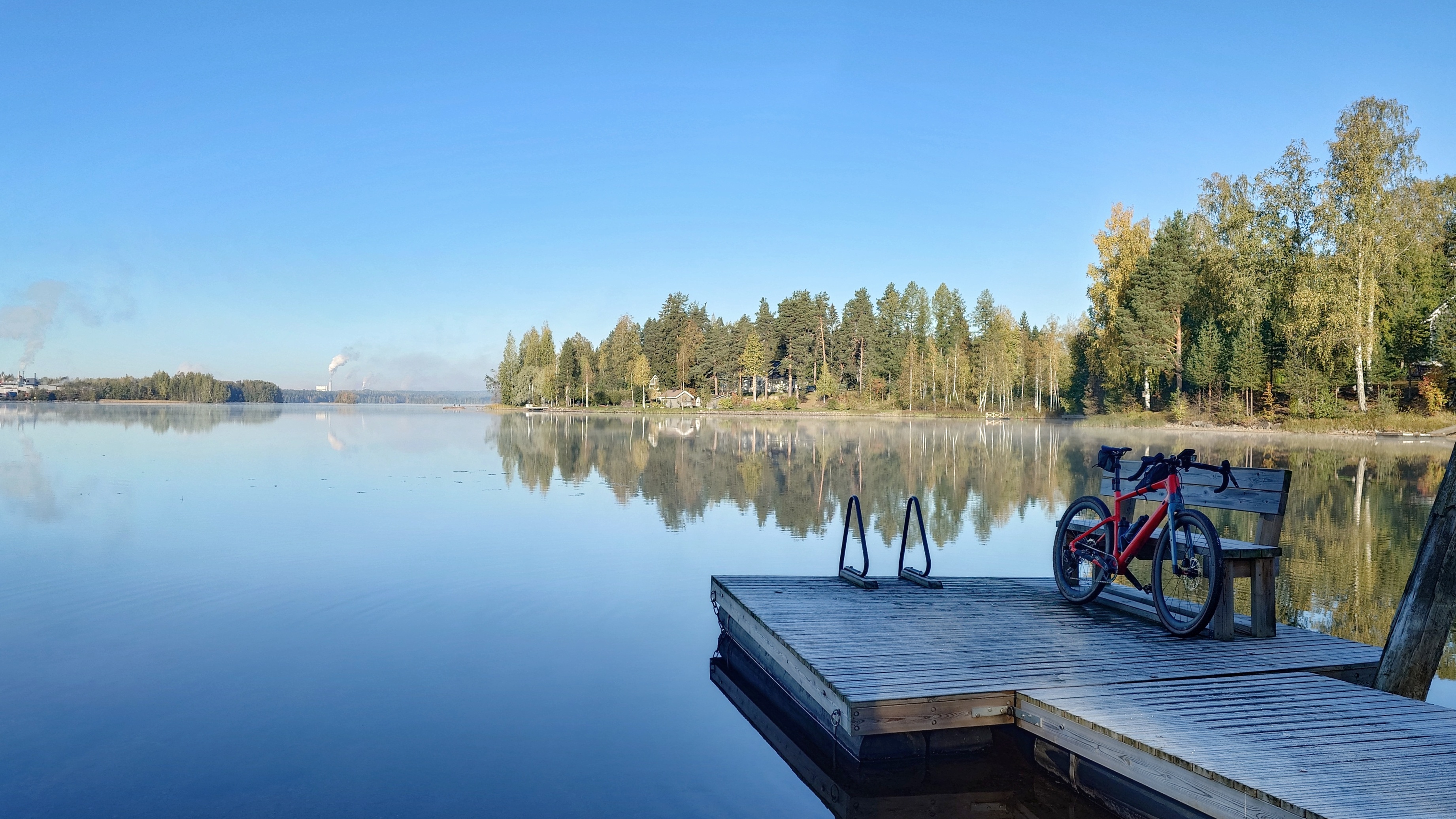 Laituri, jonka nokassa on pyörä, ympärillä oleva vesi on tyyni ja kaukana vastarannalla tehdas.