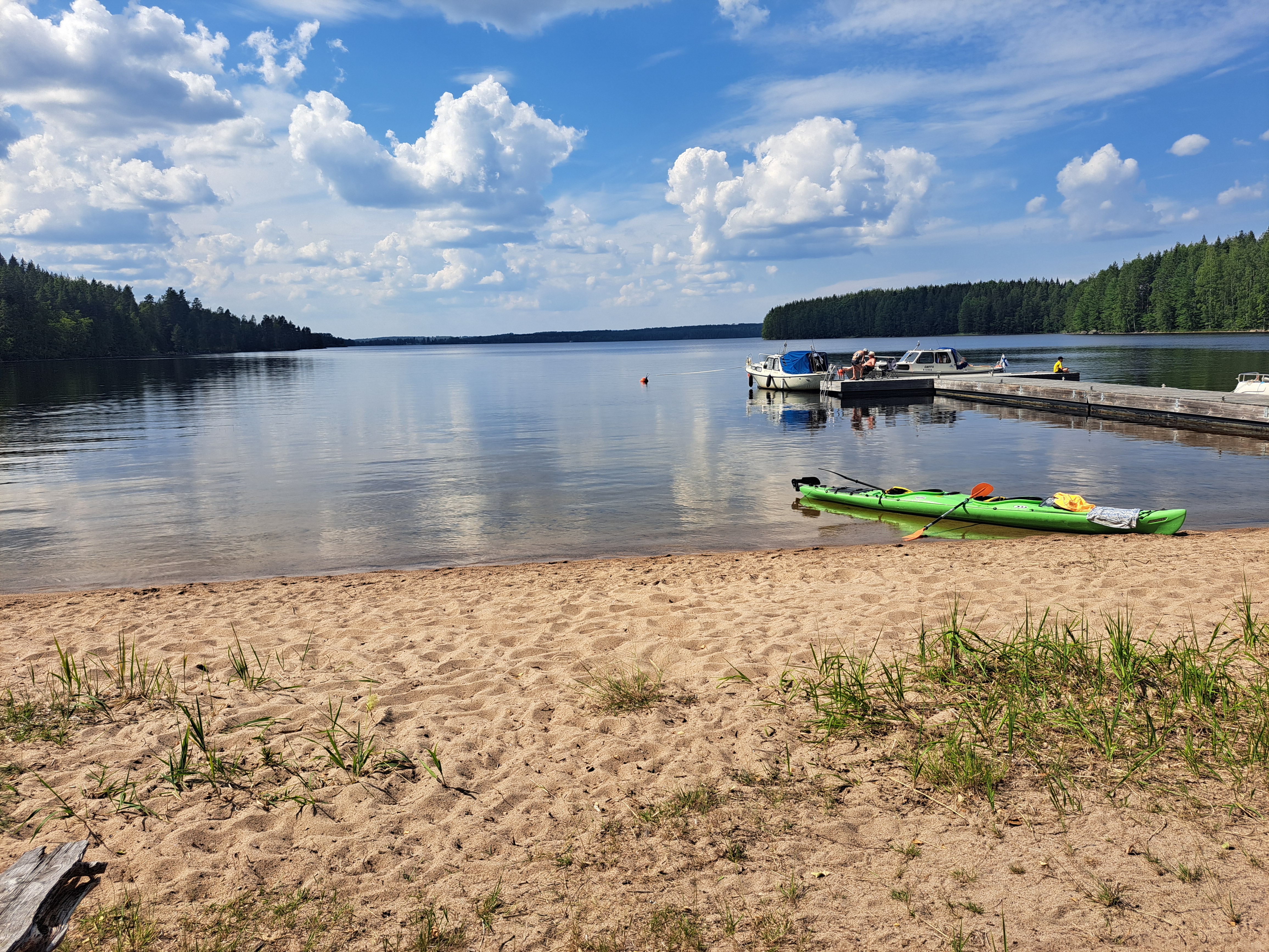 Kajakki on hiekkarannalla, ja taustalla näkyy laituri, jossa on veneitä tyynellä vedellä.