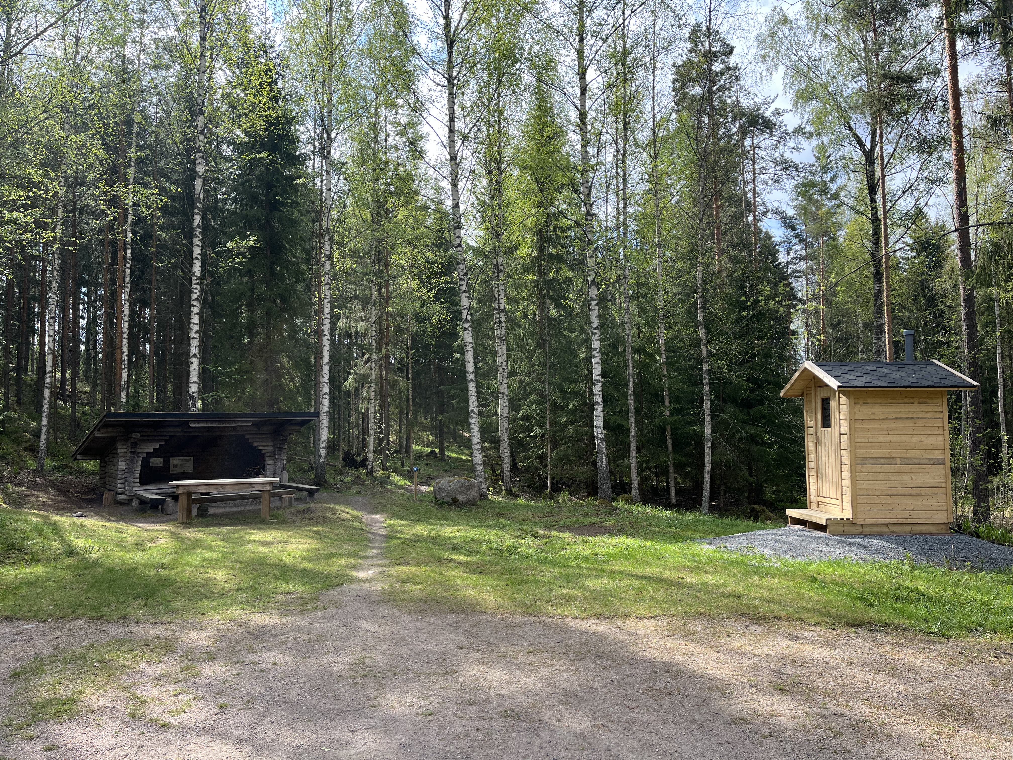 Kuvan oikeassa reunassa kuivakäymälä ja vasemmassa reunassa laavu, jonka edessä on nuotiopaikka, penkkejä ja iso puinen pöytä. Kuivakäymälän ja laavun välistä kulkee polku metsään.
