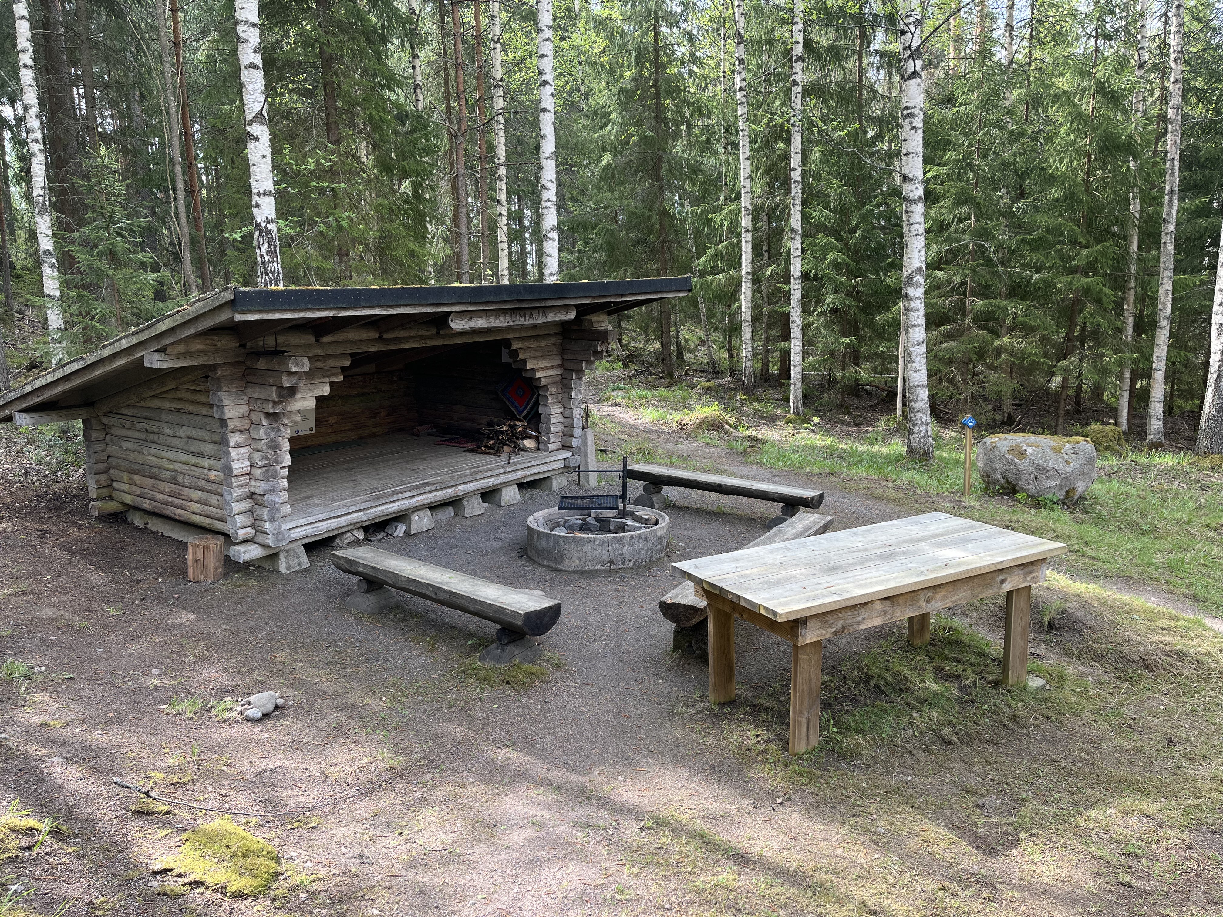 Laavu, jonka edessä on nuotiopaikka ja penkkejä kehässä. Penkkikehän ulkopuolella iso puinen pöytä.