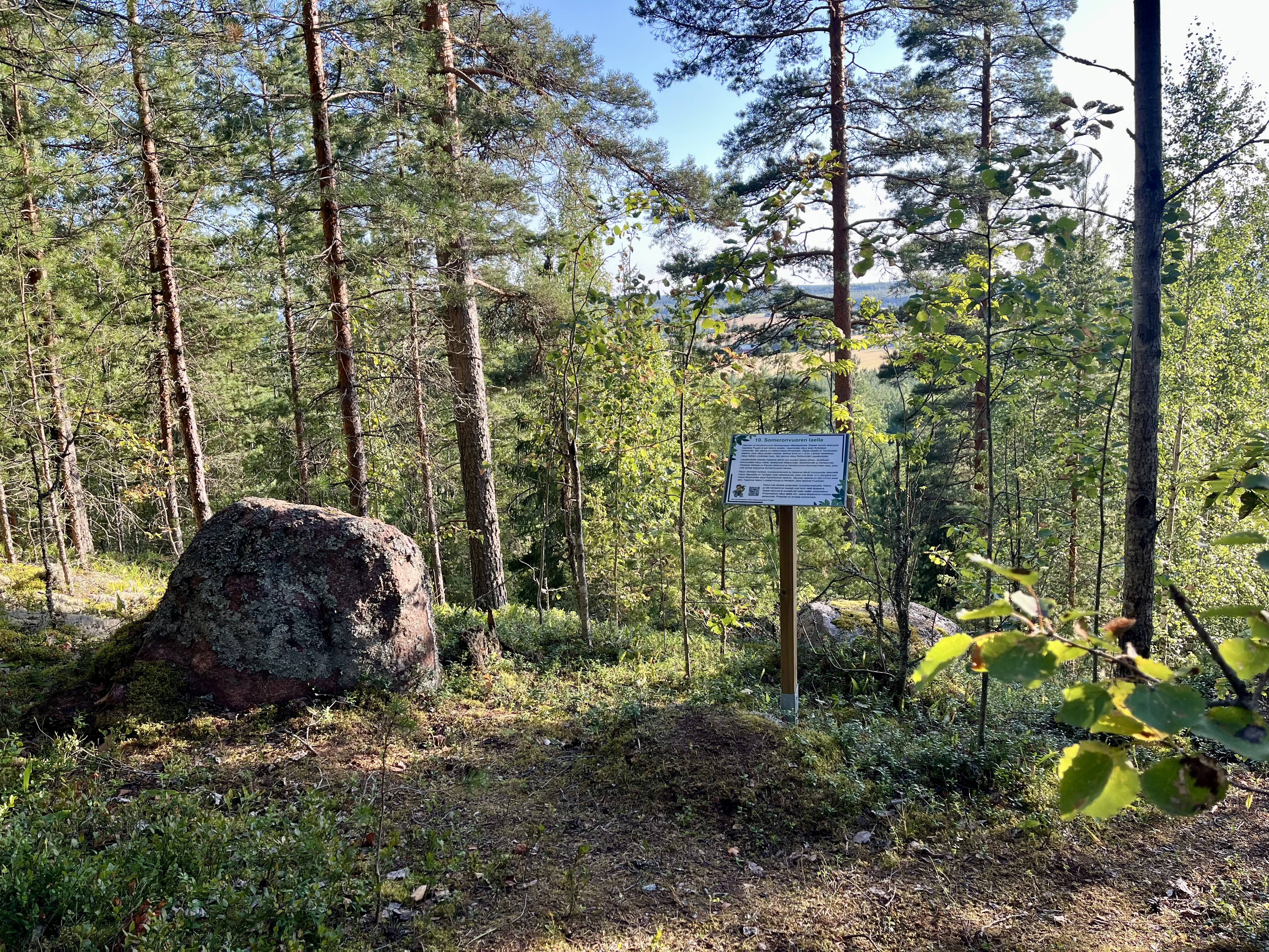 Luontopolun rastitaulu ison kiven vieressä. Taustalla puita ja maisemaa harjun laelta katsottuna.