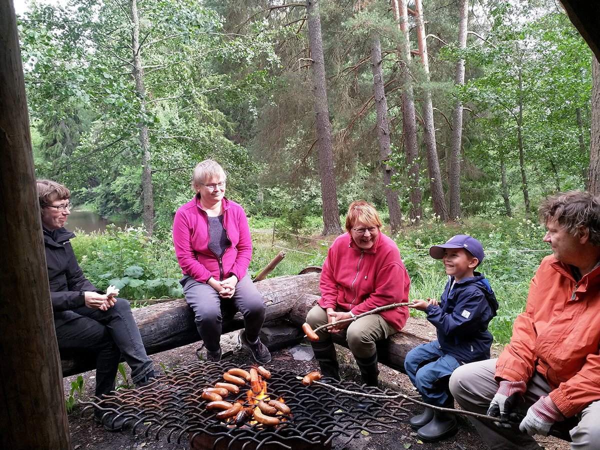 Viisi henkilöä istuu Elonkierron laavun nuotiopaikalla paistamassa makkaroita avotulella. Taustalla näkyy vehreää metsää ja Loimijoki.